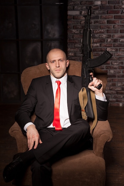 Assassino de terno e gravata vermelha sentado em uma cadeira e segurando uma arma automática