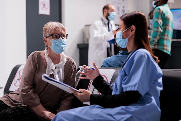 Assistente asiático consultando velha na sala de espera, falando sobre prevenção de vírus e saúde na recepção do hospital. Paciente idoso e assistente médico conversando durante a pandemia de covid 19.