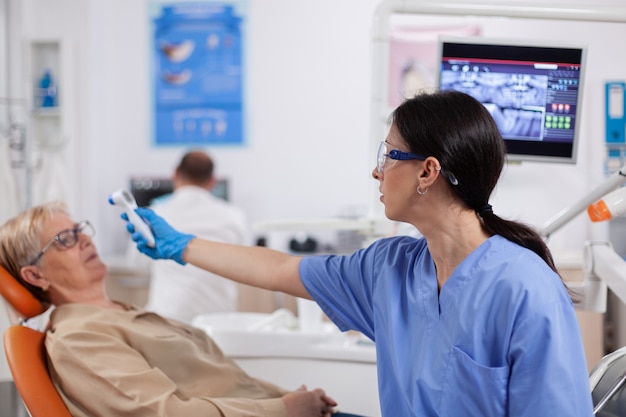 Assistente de dentista medindo a temperatura corporal da mulher sênior usando termômetro durante ...