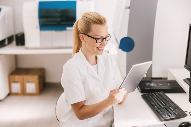 Assistente de laboratório em uniforme estéril com óculos sentado e usando um tablet para inserir os resultados do teste