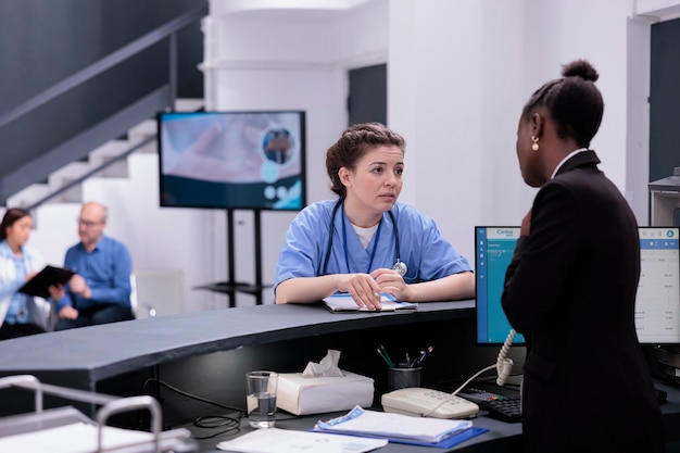 Foto assistente de pé na recepção discutindo com a recepcionista durante a consulta de check-up na sala de espera do hospital. mulheres falando sobre saúde são serviço, trabalhando para oferecer atendimento de qualidade