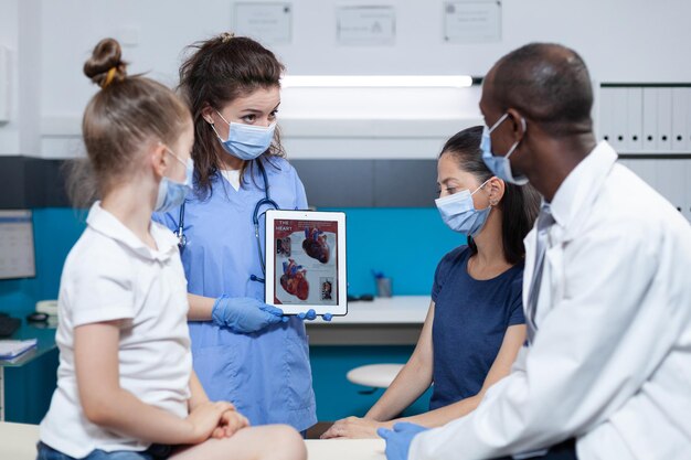 Assistente médico segurando o tablet com experiência em coração na tela durante a consulta clínica no consultório do hospital. Médico pediatra afro-americano com máscara protetora contra coronavírus