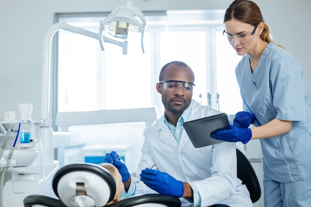 Assistente prestativo. encantadora jovem enfermeira mostrando um aviso sobre a próxima consulta no tablet para o dentista realizando um exame de cavidade oral