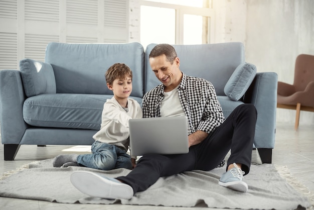 Assistindo um filme. Rapaz alegre e alegre, de cabelos louros, sorrindo e sentado no chão com o pai e eles usando o laptop