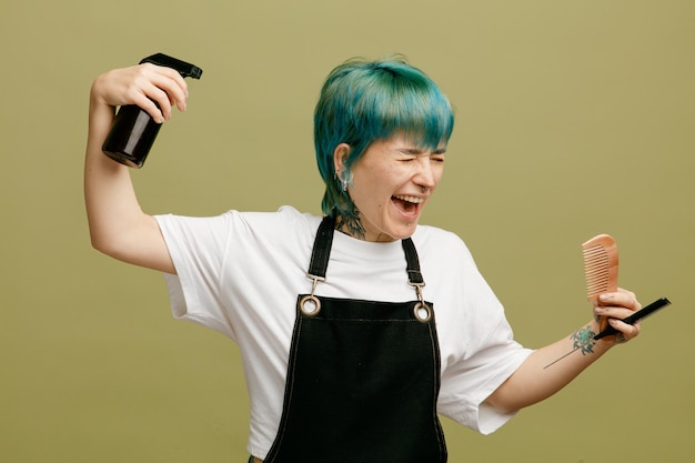 Assustado jovem barbeiro vestindo uniforme segurando pentes aplicando spray de cabelo no cabelo dela virando a cabeça para o lado gritando com os olhos fechados isolados no fundo verde-oliva