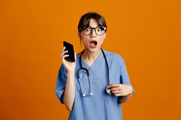 assustado segurando o telefone jovem médica usando estetoscópio uniforme de quinto isolado em fundo laranja