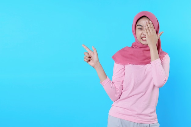 Assustar a mulher asiática com o dedo apontando isolado em um fundo azul claro usando hijab com camiseta rosa