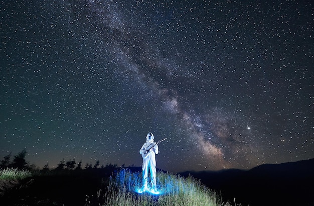 Astronauta iluminado fresco do espaço parado no meio do prado de montanha segurando guitarra sob a Via Láctea