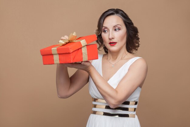 Astuta mulher morena elegante de meia-idade em vestido branco segurando e olhando para dentro da caixa de presente vermelha. conceito de emoção e sentimento. foto de estúdio, interna, isolada em fundo marrom claro