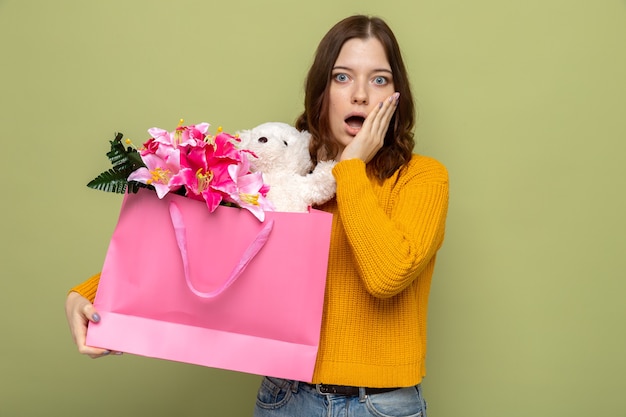 Asustada poniendo la mano en la mejilla hermosa joven sosteniendo una bolsa de regalo