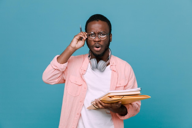 Asustado sosteniendo libros con bolígrafo joven afroamericano con auriculares en el cuello aislado de fondo azul