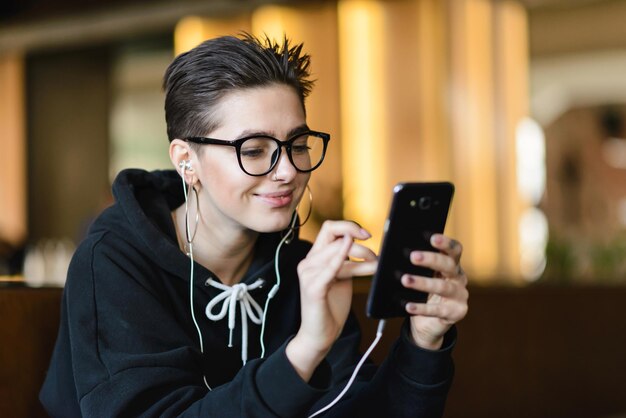 Atalho hipster jovem assistindo vídeos engraçados no smartphone através de fones de ouvido modernos sentados no café