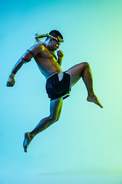 Ataque. Boxeador tailandés joven posando sobre fondo azul en luz de neón. Luchador practicando, entrenando en artes marciales en acción. Salud, deporte, concepto de cultura asiática. Copie el espacio para el anuncio. Muay thai.