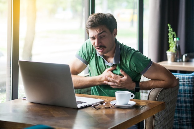 Ataque cardíaco Jovem empresário em camiseta verde sentado e segurando seu peito porque sentindo dor em seu negócio de coração e conceito de freelancing tiro interno perto de janela grande durante o dia