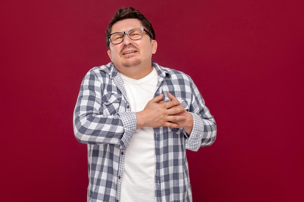 Ataque de dolor al corazón o infarto. Retrato de un hombre de mediana edad con camisa a cuadros casual y anteojos de pie y sosteniendo su doloroso corazón. tiro de estudio interior, aislado sobre fondo rojo oscuro.