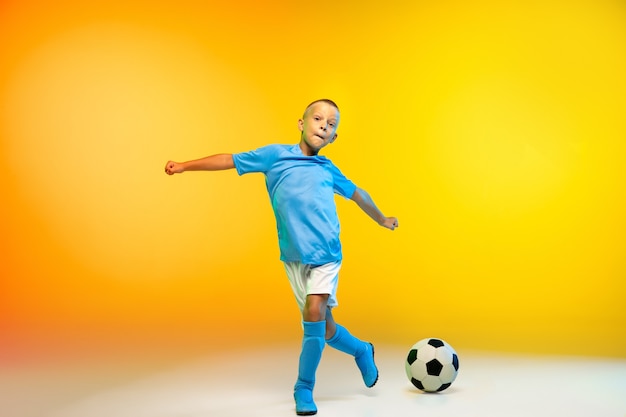 Ataque. Joven como jugador de fútbol o fútbol en ropa deportiva practicando sobre fondo de estudio amarillo degradado en luz de neón. Encajar jugando al niño en acción, movimiento, movimiento en el juego. Copyspace.