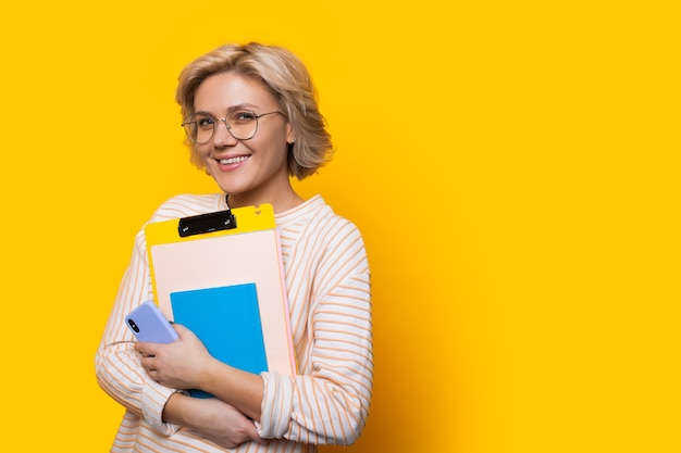 Atemberaubende blonde Lehrerin, die durch Brillen schaut, während sie einige Bücher hält und auf einer gelben Wand mit freiem Raum aufwirft