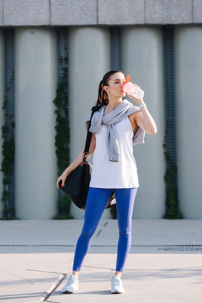 Atemberaubende Frau in sportlichem Outfit, die auf der Straße Trinkwasser steht