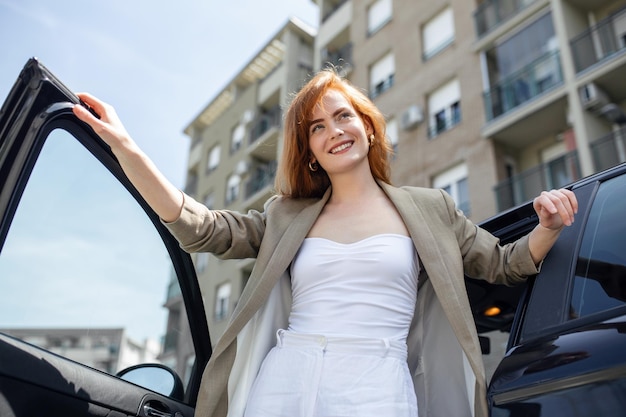 Atemberaubende junge Frau beim Betreten ihres Autos, die mit einer Hand die Tür hält und sich mit der anderen auf die Motorhaube stützt