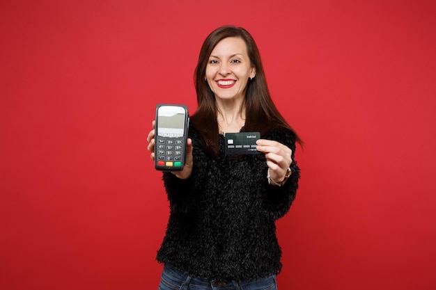 Atemberaubende junge Frau in schwarzem Pelzpullover hält drahtloses modernes Bankzahlungsterminal, um Kreditkartenzahlungen einzeln auf rotem Hintergrund zu verarbeiten und zu erwerben. Menschen aufrichtige Emotionen, Lifestyle-Konzept.