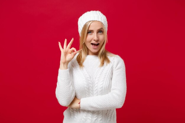 Foto atemberaubende junge frau in weißem pullover, hut isoliert auf rotem hintergrund, studioporträt. gesunder modelebensstil, menschen aufrichtige emotionen, konzept der kalten jahreszeit. mock-up-kopienbereich. ok-geste wird angezeigt.