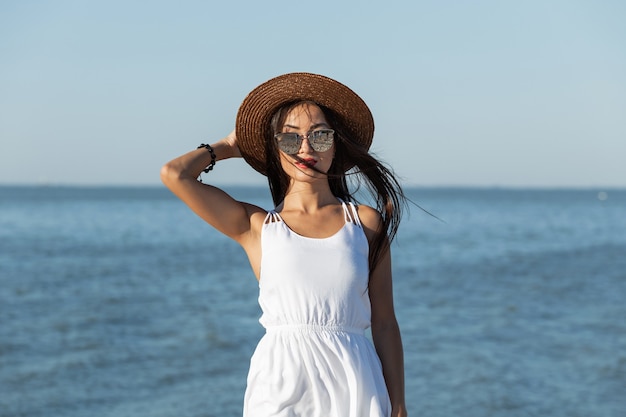 Atemberaubendes dunkelhaariges Mädchen in weißem Kleid, Sonnenbrille und braunem Hut am Meer an einem sonnigen Tag. .