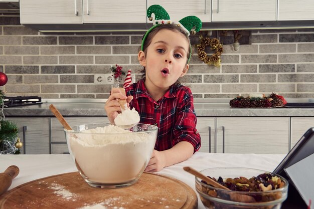 Atemberaubendes positives kaukasisches kleines Mädchen, das Weihnachtskekse und Brot in der heimischen Küche kocht, schaut in die Kamera, die einen Holzlöffel voller Weißmehl in ihrer ausgestreckten Hand zeigt