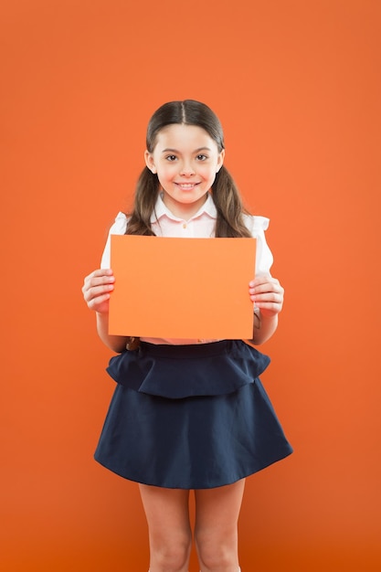 Atenção, por favor, menina da escola, segure papel no espaço de cópia de fundo laranja menina feliz em uniforme escolar com informações úteis de volta à escola, olhe aqui escrevendo notas educação moderna