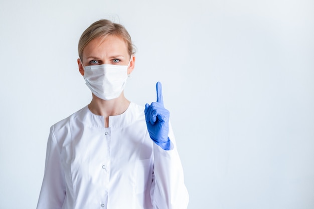 Atención. Doctora rubia cirujano en uniforme blanco, guantes protectores azules y máscara médica mostrando el dedo hacia arriba sobre gris.