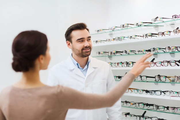 atención de la salud, personas, vista y concepto de visión - mujer feliz eligiendo y mostrando anteojos al óptico en la tienda de óptica