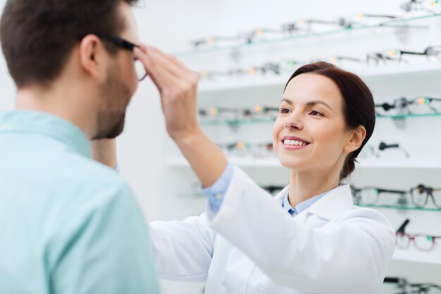atención de la salud, personas, vista y concepto de visión - óptica femenina poniéndose anteojos al hombre en la tienda de óptica