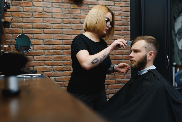 Atendendo cliente na Barbearia. Barbeiro profissional, cabeleireira feminina fazendo corte de cabelo moderno para um homem sentado na cadeira da barbearia.