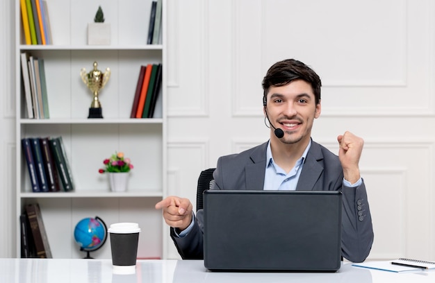 Atendimento ao cliente bonitinho de terno cinza com computador e fone de ouvido apontando para o copo de papel