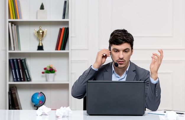 Atendimento ao cliente bonitinho de terno cinza com computador e fone de ouvido com papéis espremidos na mesa