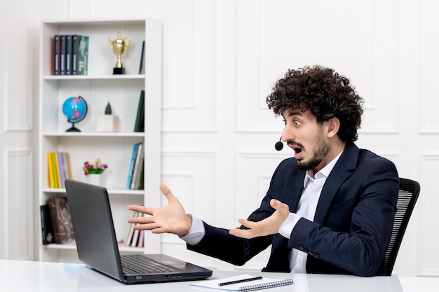 Atendimento ao cliente bonito homem encaracolado em terno de escritório com computador e fone de ouvido chocado e confuso