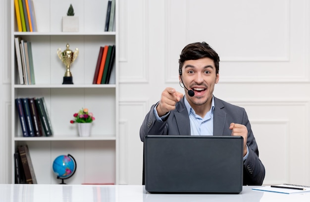 Atendimento ao cliente homem bonito de terno cinza com computador e fone de ouvido apontando para a frente