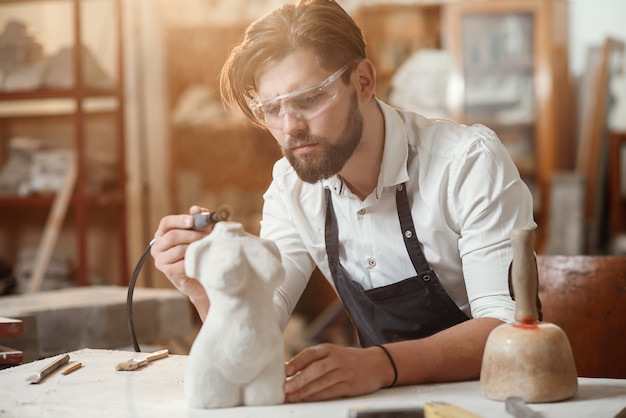 Atento albañil con elegante barba en gafas protectoras que detalla la escultura creativa del torso de la mujer en su lugar de trabajo.