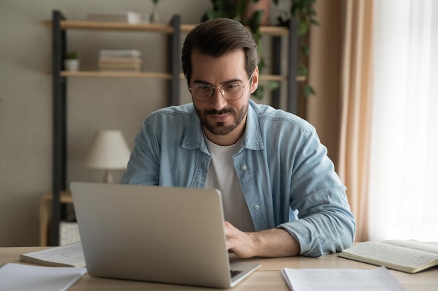 Atento gerente masculino da geração do milênio ocupado em teletrabalho no tempo de quarentena estudando documentos na rede corporativa de casa PC jovem especialista concentrado fazendo trabalho de computador remoto em laptop