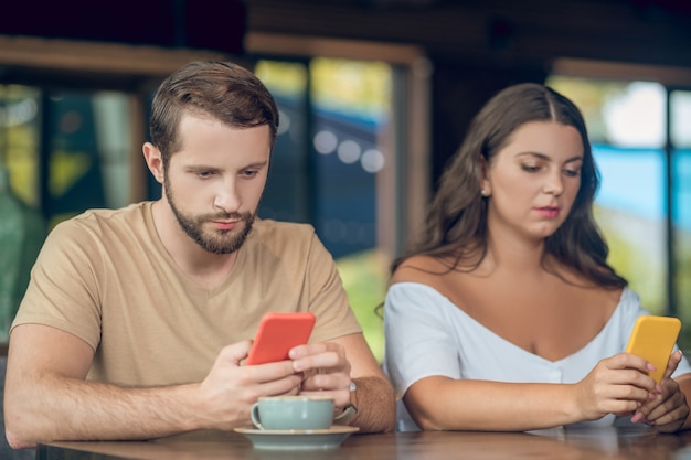 Atento joven y mujer mirando fijamente a los teléfonos inteligentes mientras está sentado con un café en la cafetería