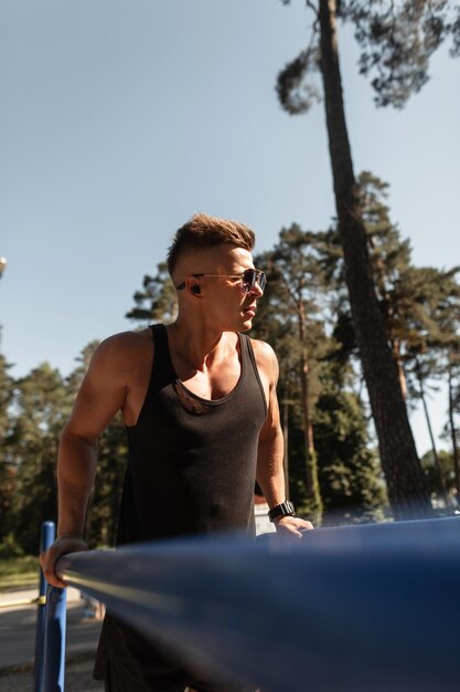 Athlet gutaussehender Mann mit Sonnenbrille und kabellosen Kopfhörern in schwarzen Kleidern Liegestütze auf Bars und Musik im Freien hören