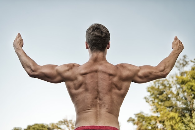 Athlet mit nacktem Oberkörper am grauen Himmel. Bodybuilder, der Muskeln, Bizeps und Trizeps zeigt. Mann oder Sportler, die Arme anheben, Rückansicht. Fitness und Sport. Gesundes Lebensstilkonzept