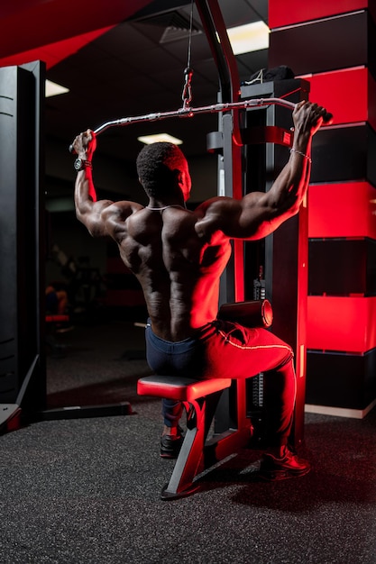 Athlet pumpt Bizeps auf dem Simulator. Afrikanischer gutaussehender Bodybuilder im Fitnessstudio. Ansicht von hinten.