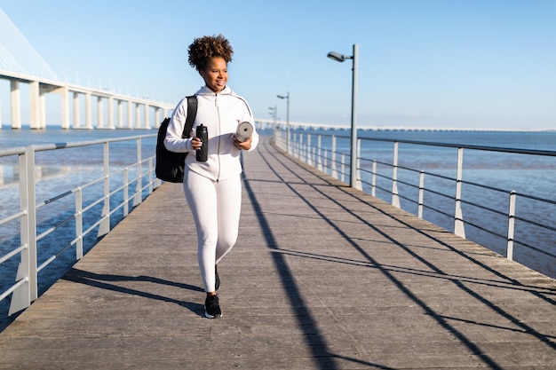 Athletische afrikanisch-amerikanische Dame, die mit Fitnessmatte und Wasserflasche im Freien spazieren geht