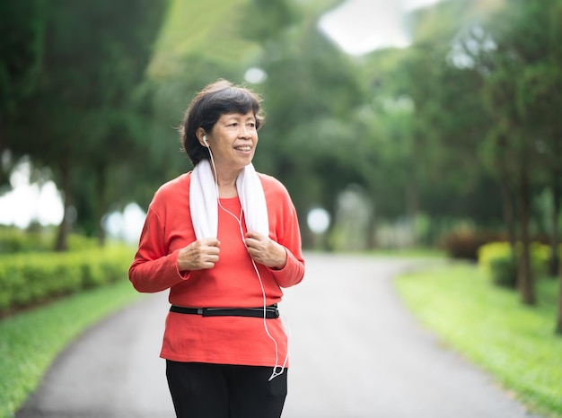 Athletische asiatische Seniorin der 60er Jahre, die lächelt und joggt Schöne asiatische Seniorin, die an einem sonnigen Tag im Park läuft Läuferin, die beim Joggen Musik hört