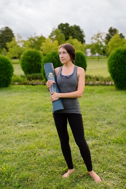 Athletische dünne junge Frau hält nach oder vor dem Training im Freien eine Yogamatte und eine Flasche Wasser.