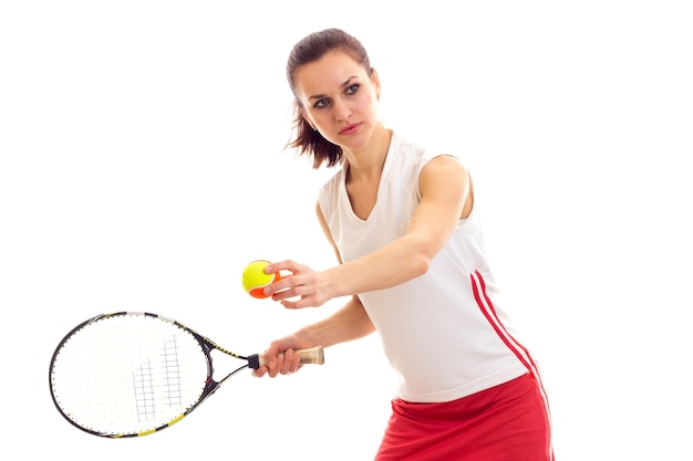 Athletische, timistische junge Frau in weißem Sporthemd und rotem Rock mit kastanienbraunem Pferdeschwanz mit Tennisschläger und gelbem Ball auf weißem Hintergrund im Studio