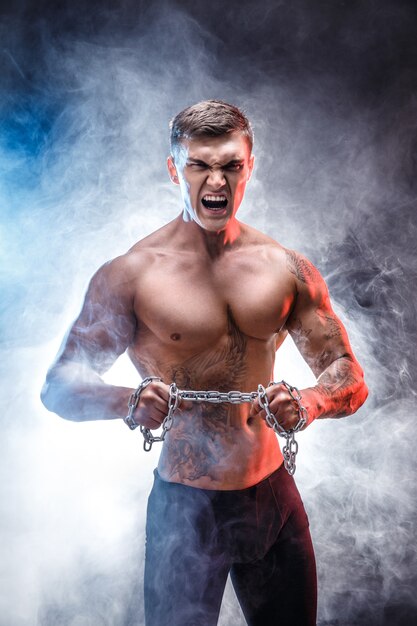 Athletischer Mannbodybuilder der hübschen Energie, der Übungen mit der Kette, zerreißend tut. Muskulöser Körper der Eignung auf dunklem Hintergrund. Perfekter Mann. Fantastischer Bodybuilder, Tätowierung, werfend auf.