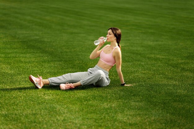 Athletisches junges Mädchen trinkt Wasser nach einem harten Training, das im Sommer auf dem Gras sitzt