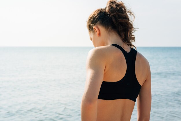 Athletisches Mädchen mit dem gelockten Haar in einem Sport-BH, der auf dem Ufer steht und das Wasser bei Sonnenaufgang betrachtet