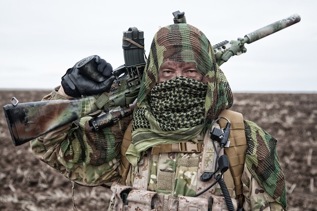 Foto atirador do exército, jogador de airsoft com uniforme camuflado e carregador de carga, capa de máscara na cabeça, rifle de serviço armado com mira óptica, escondendo rosto com shemagh, em pé no campo, olhando para longe